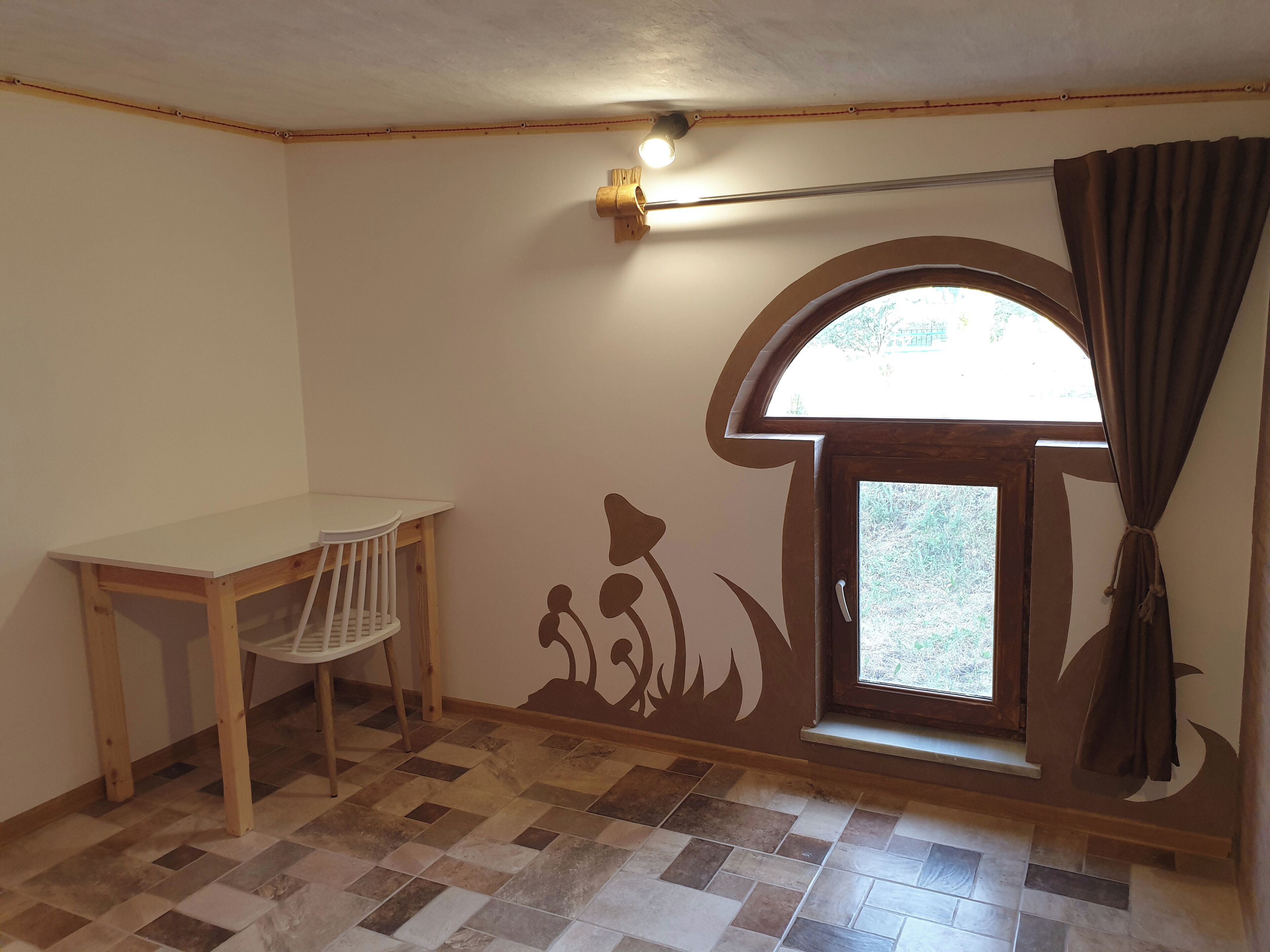 A room with a wooden desk, a chair, a window with curtains, and a wall mural.