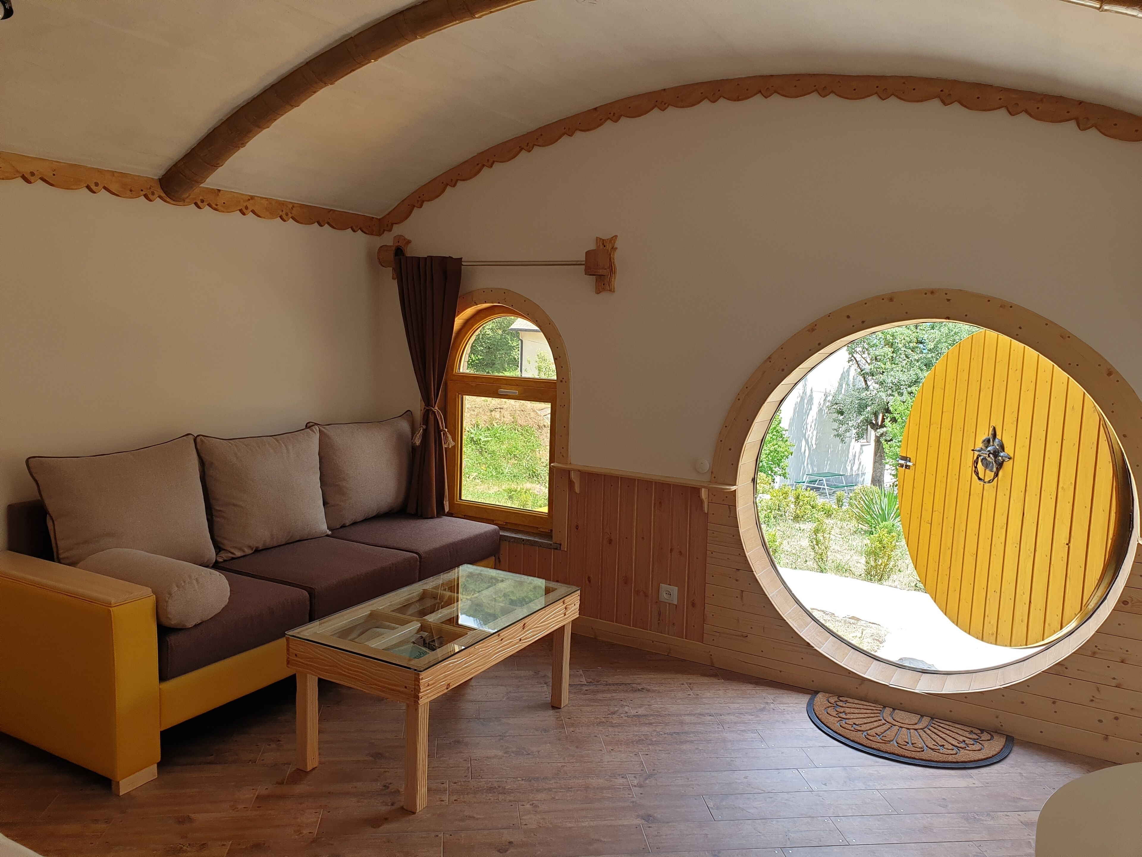 Une pièce chaleureuse avec un canapé en bois, une table basse en verre et une grande fenêtre circulaire surmontée d’une porte jaune.
