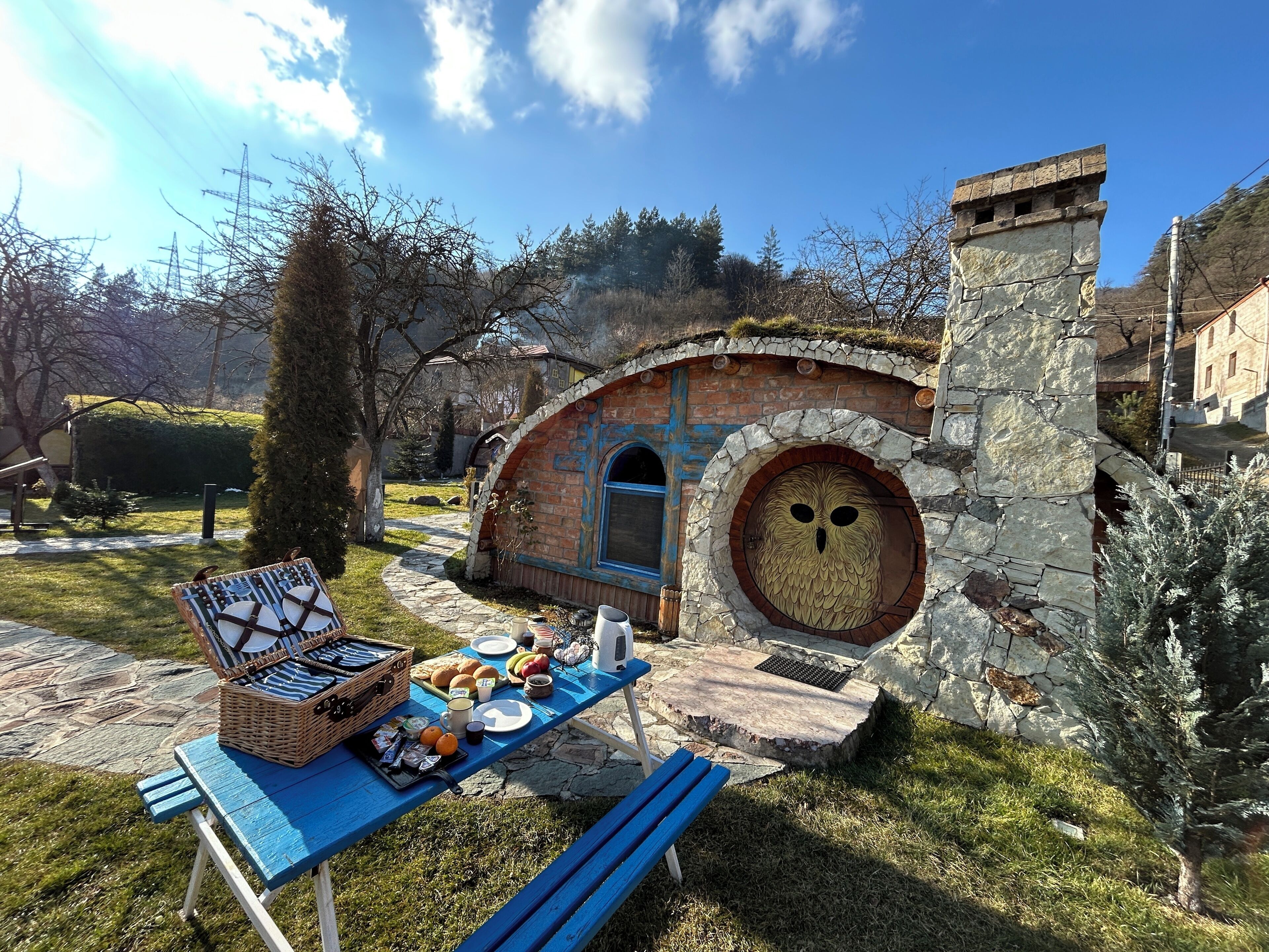 Un comedor rústico al aire libre con un horno de piedra y una mesa azul dispuesta con comida y bebidas.
