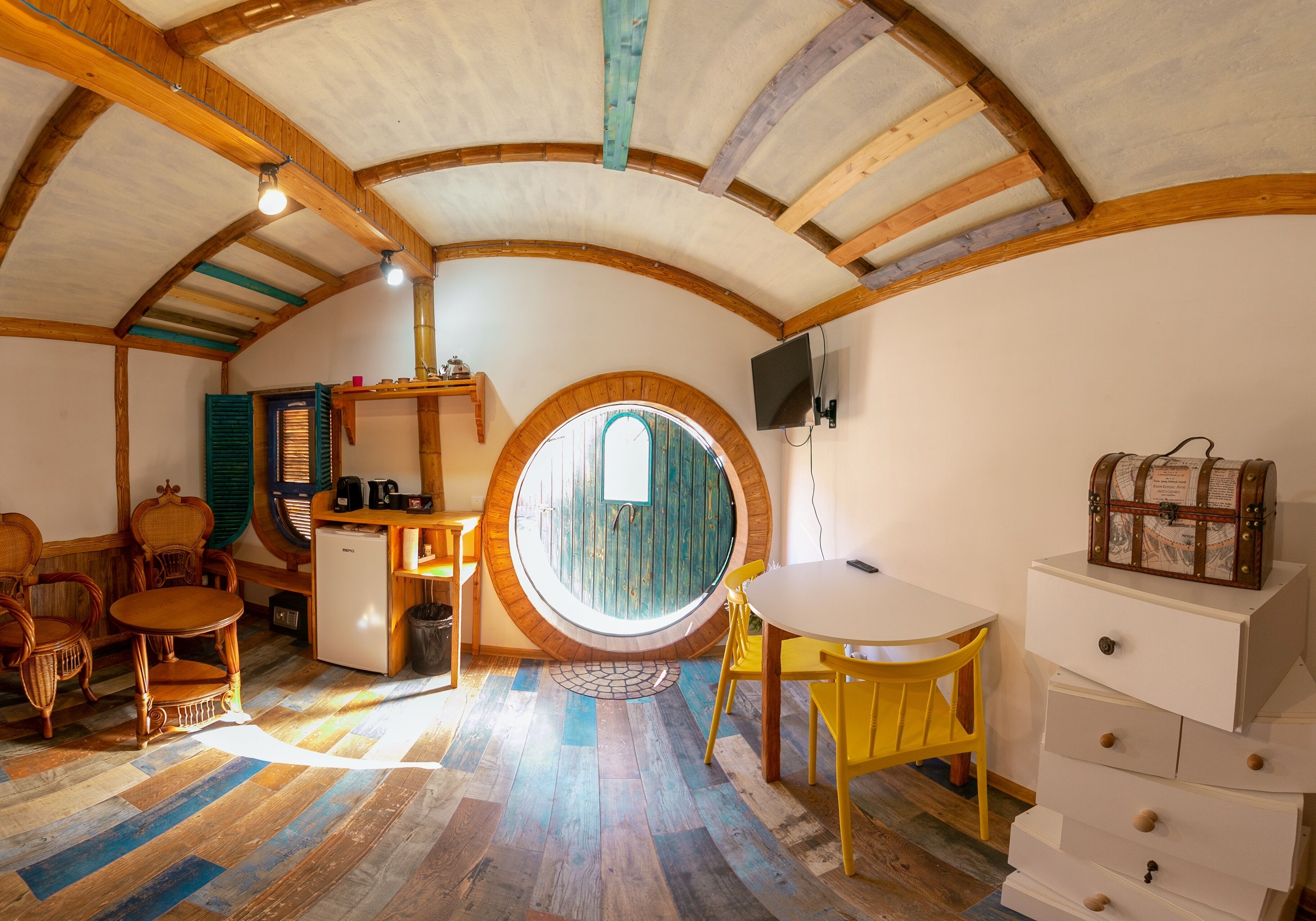 A cozy, circular room with wooden beams, a round window, a small table, chairs, a desk, and a dresser.