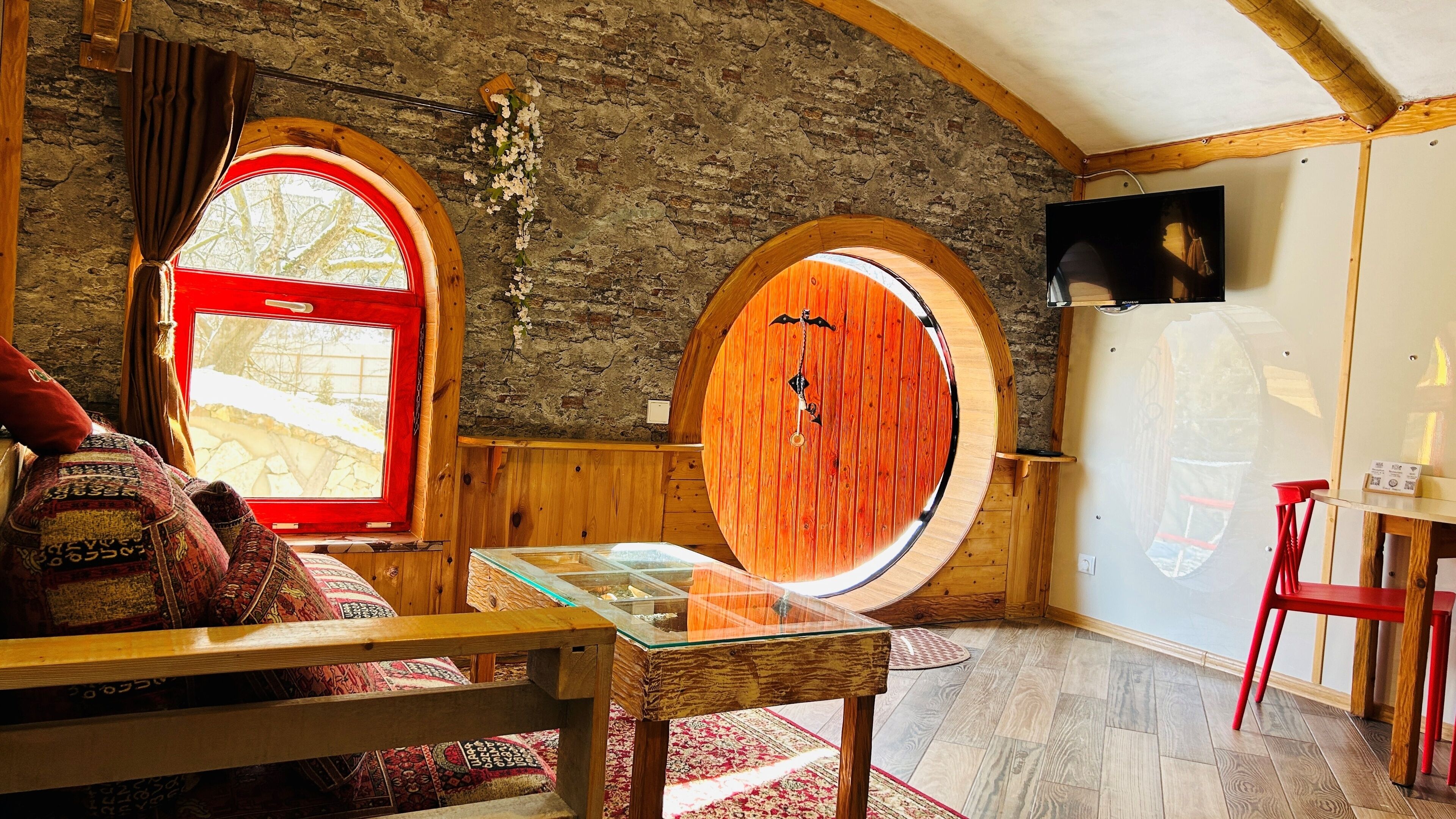 A cozy room with a wooden ceiling, a large window with a red frame, and a circular wooden door.