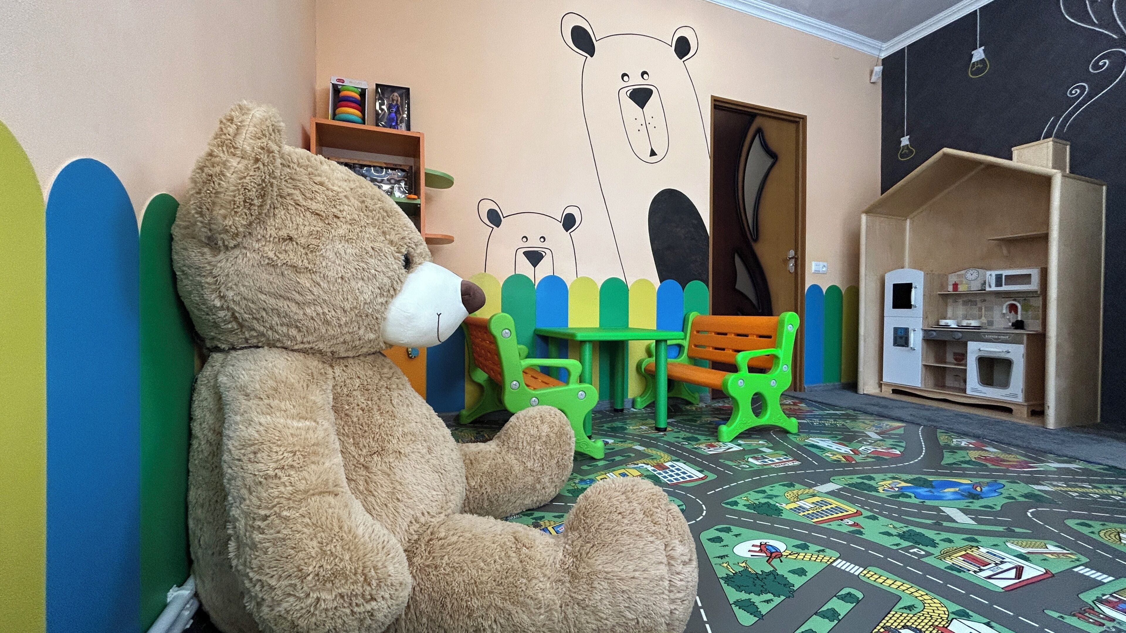 A room with a large teddy bear, a green and orange play table, and a small kitchen area.