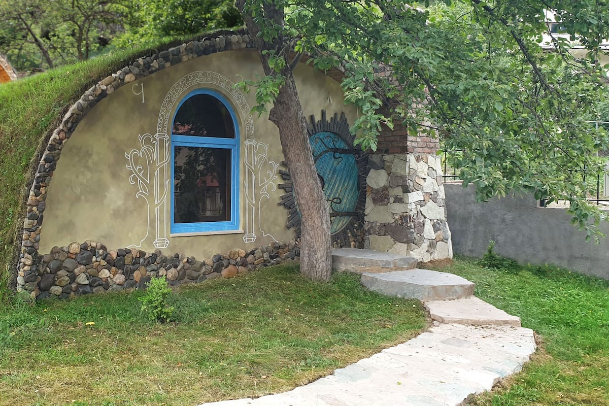 A small, stone-walled structure with a blue window and a decorative archway, surrounded by grass and trees.