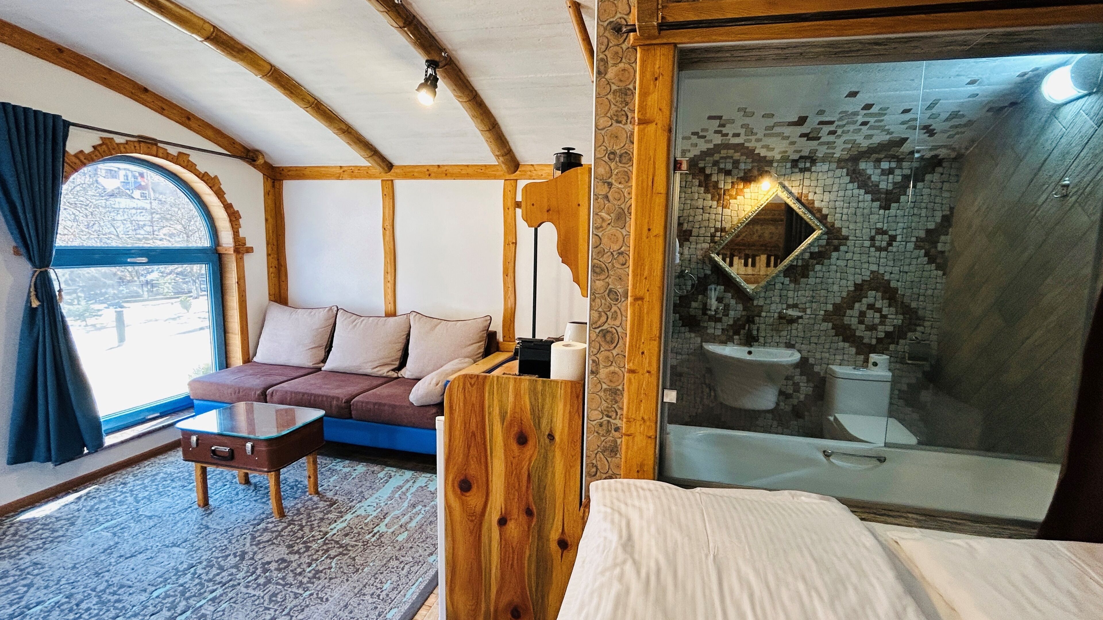 A cozy room with a wooden ceiling, a sofa, a small table, and a bed. There's a window with curtains and a view outside.
