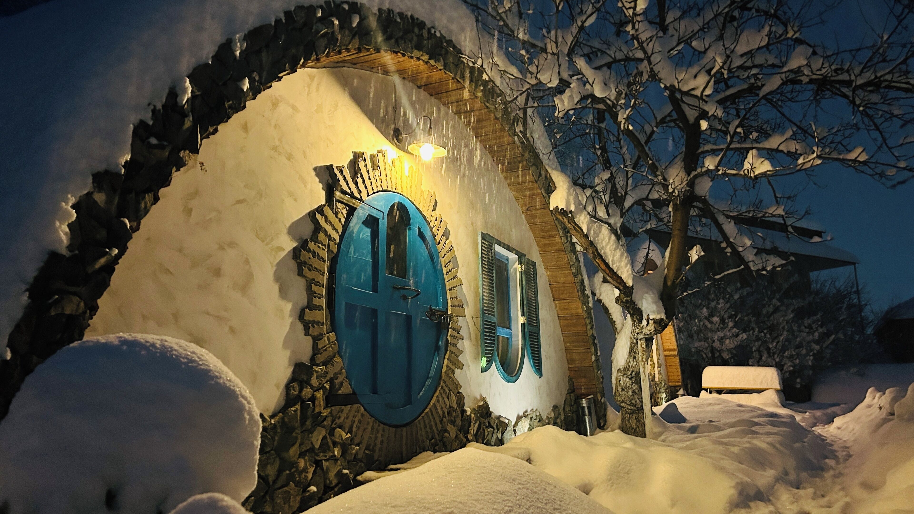 A snow-covered building with a distinctive blue door and windows, surrounded by snow and trees.