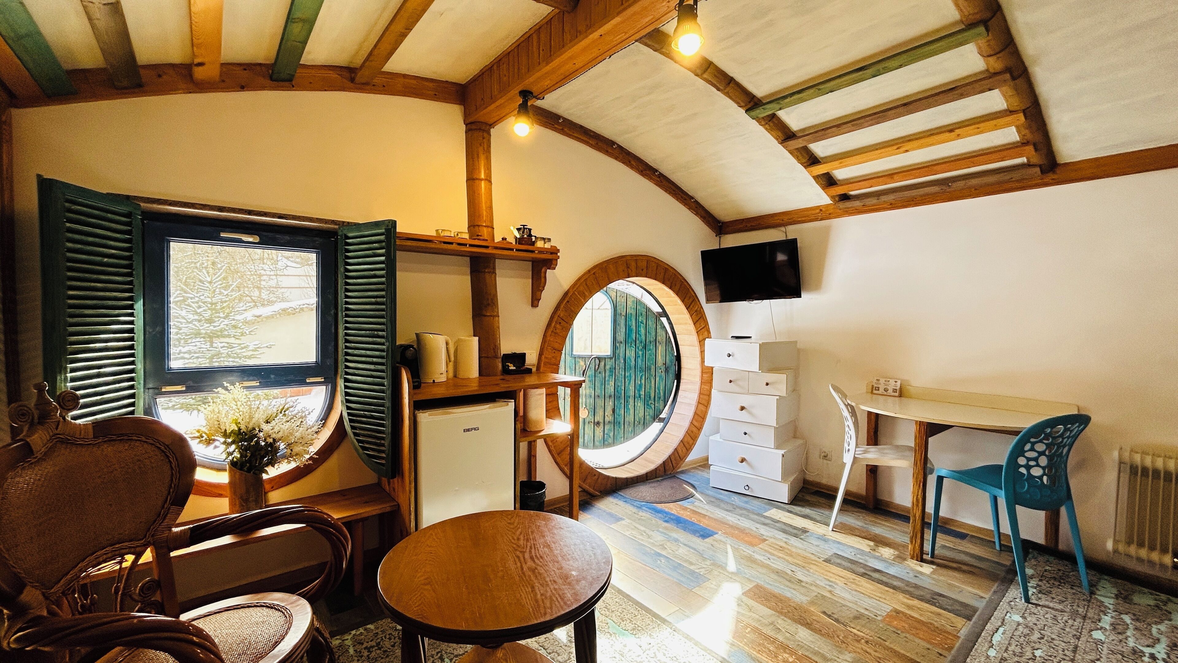 A cozy room with a wooden ceiling, a round window, a desk, a chair, and a television.