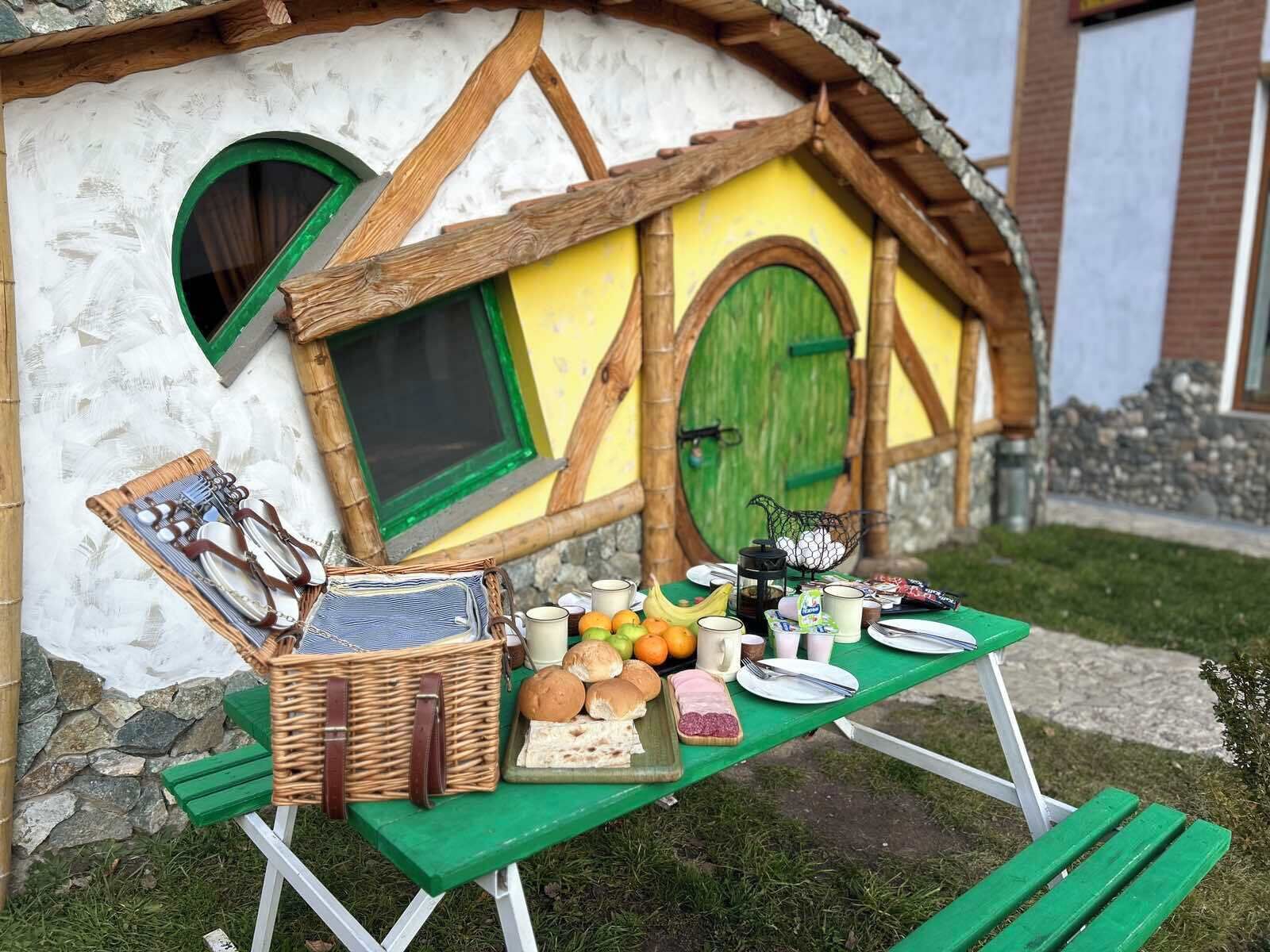 Une petite maison avec une porte et des fenêtres vertes, une table de pique-nique avec de la nourriture et des boissons, et un panier en osier.
