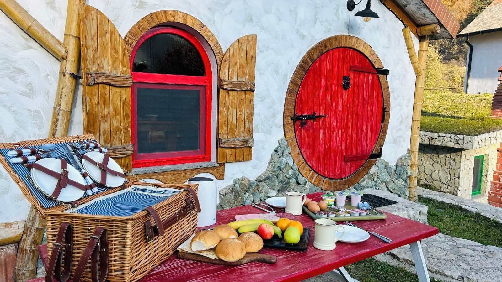A rustic outdoor dining setup with a wicker basket, plates, and a variety of fruits and bread on a red table.