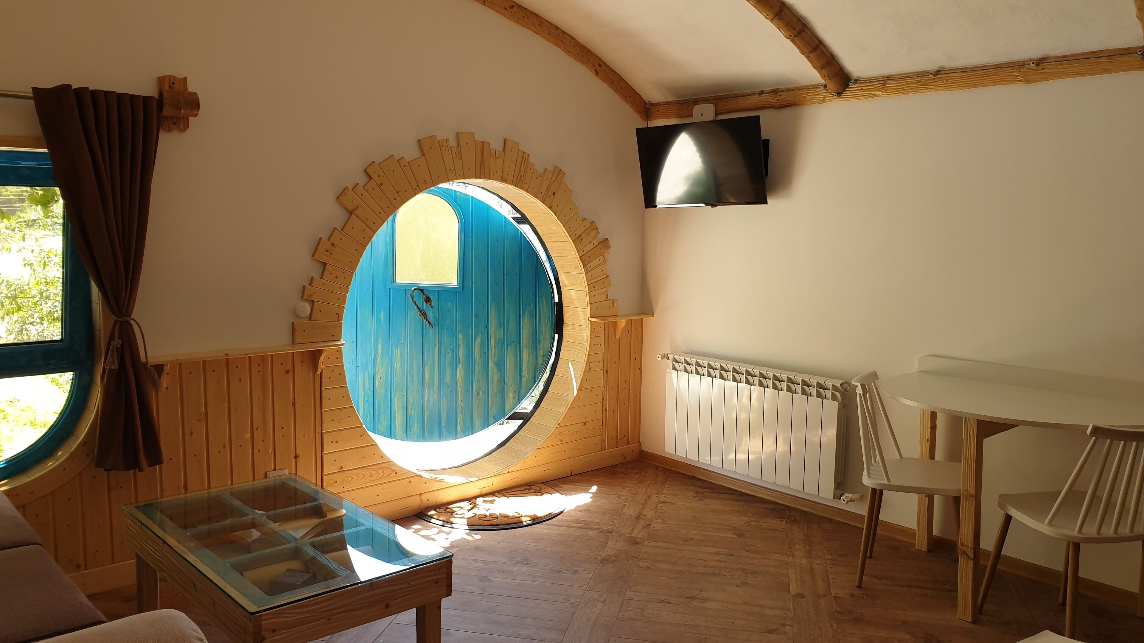 A cozy room with a wooden ceiling, a circular window with a blue door, a glass display table, and a white chair.
