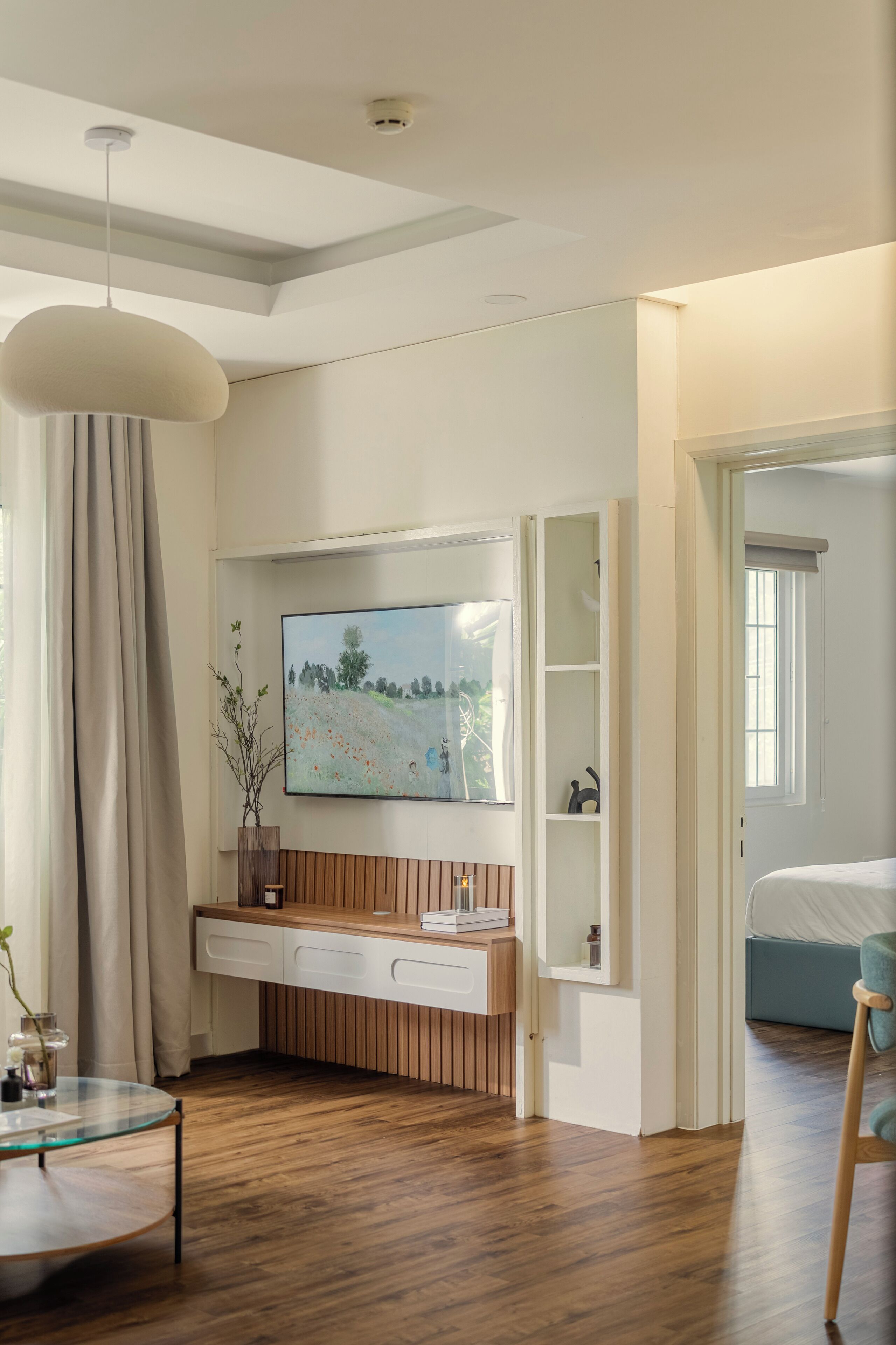 A modern bedroom with a wooden floor, a glass coffee table, a bed, a TV mounted on the wall, and a window with curtains.