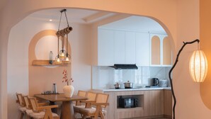 Un espace de vie et de salle à manger à aire ouverte, avec une table à manger en bois, des chaises, une petite table basse ronde sur laquelle sont posés des livres et des fruits, une cuisine en arrière-plan et un grand lampadaire.