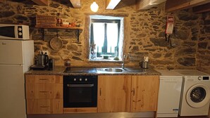 A rustic kitchen with a stone wall, wooden cabinets, a built-in oven, a washing machine, and a window with curtains.