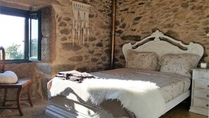 A stone-walled bedroom with a bed, a chair, a hanging macrame piece, and a window.