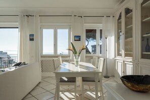 Enjoy meals with a view of the sea from this dining area, featuring a table for two, a vase with flowers, and large windows.