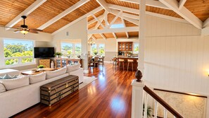 A spacious living area with a wooden ceiling, a large window, a sofa, a television, and a kitchen area with a dining table and chairs.