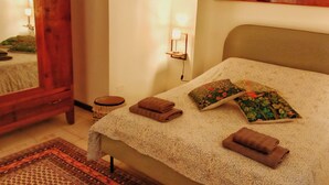 A neatly made bed with a patterned bedspread, two pillows with floral designs, and two folded towels. There is a wooden side table with a lamp, a small round stool, and a mirror on the wall. - Cozy apartment in historic country residence, between Perugia and Assisi (Perugia)