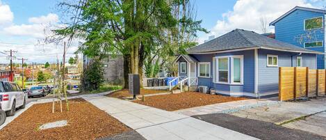 A residential area with a blue house, a sidewalk, a tree, and parked cars.