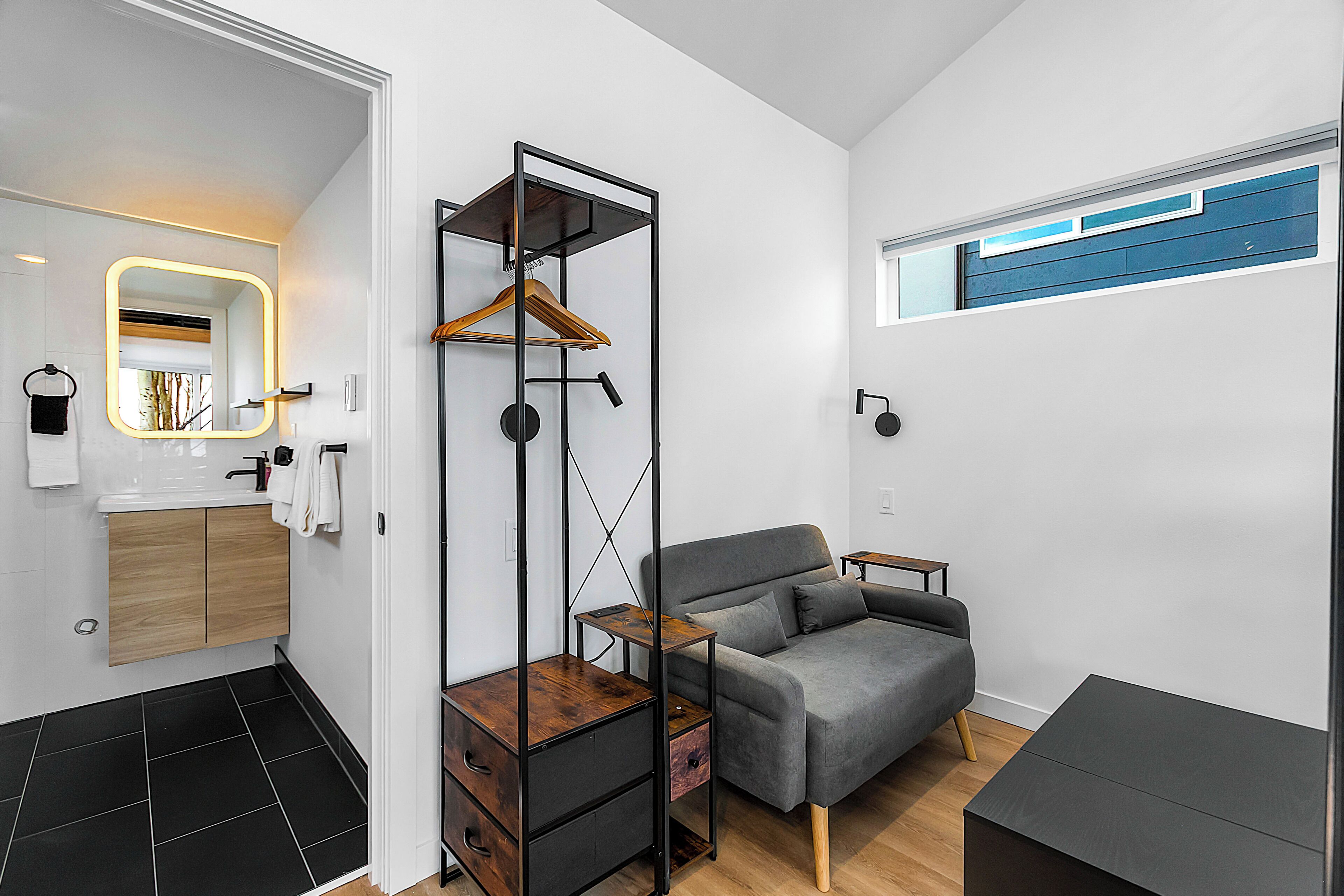 A modern, compact living space with a grey sofa, a wooden cabinet, a tall shelving unit, and a small table.