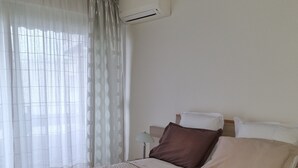 A bedroom with a bed, a nightstand, an air conditioning unit, and sheer curtains.