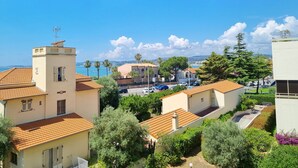 A coastal residential area with terracotta-roofed buildings, a clear blue sky, and a view of the sea in the background.