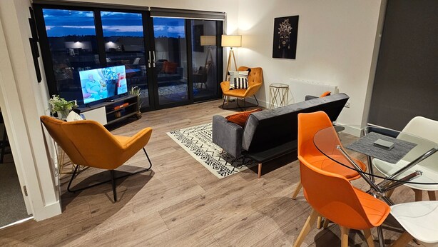 A modern living room with a large TV, orange chairs, a glass table, and a view of the city through large windows. - Balcony View Apartment- Sleeps 5 & Private Parking (Solihull)
