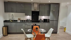 A modern kitchen with grey cabinets, a stainless steel refrigerator, and a dining area with a glass table and two chairs. - Balcony View Apartment- Sleeps 5 & Private Parking (Solihull)