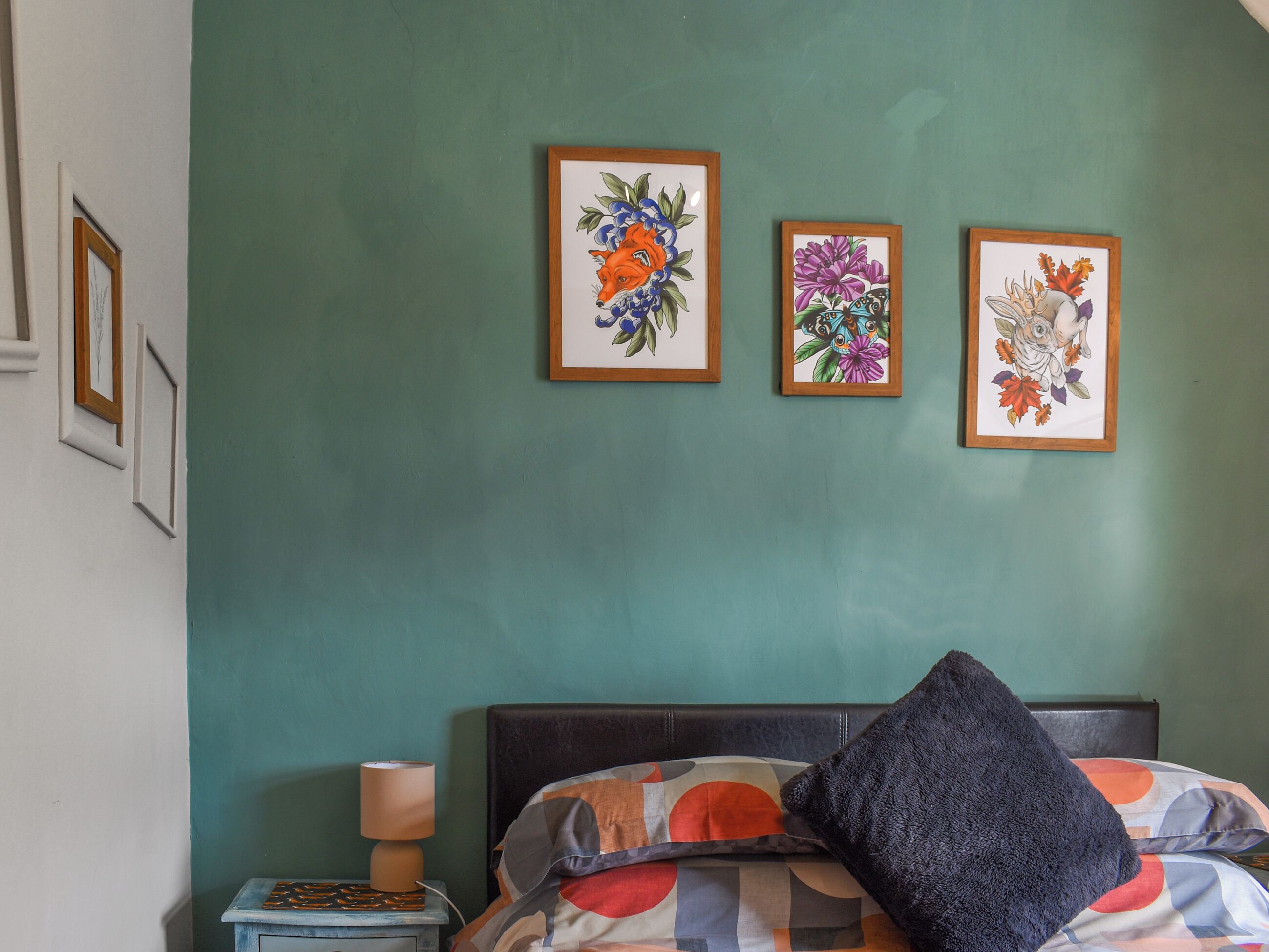 A bedroom with a bed, a nightstand, a lamp, and three framed floral artworks on the wall.