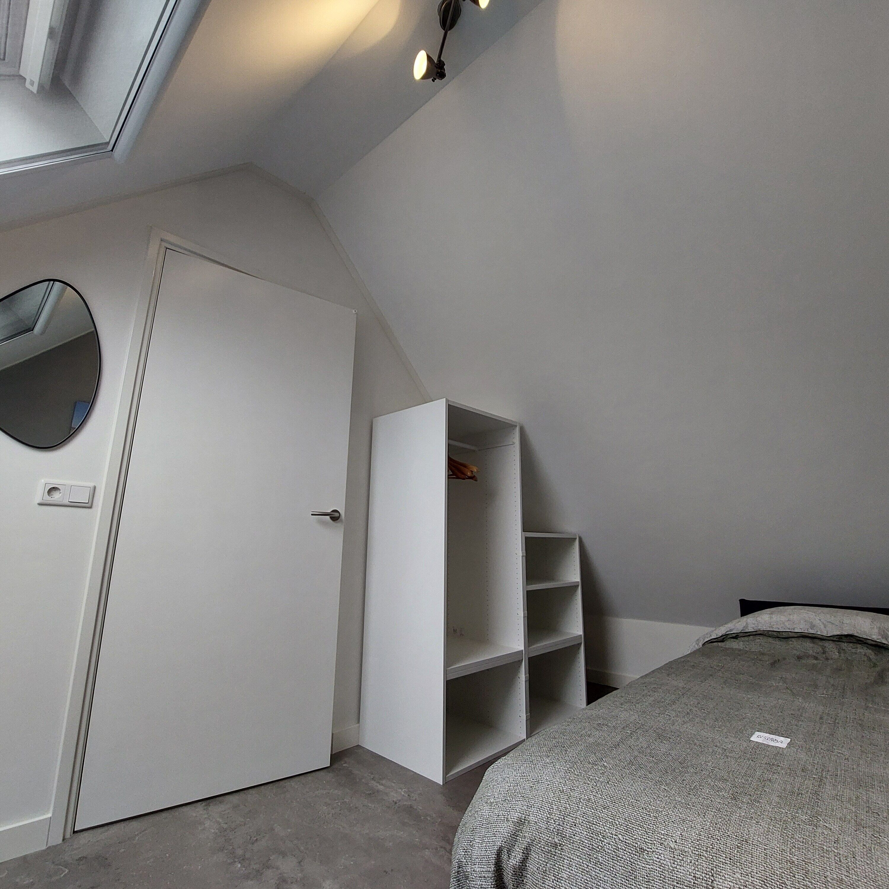 A bedroom with a white door, a built-in wardrobe, a bed, and a small circular mirror.