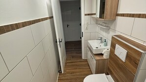 Un baño estrecho con inodoro, lavamanos y espejo. Las paredes están cubiertas de azulejos con un patrón de espiga, y el suelo es de madera.