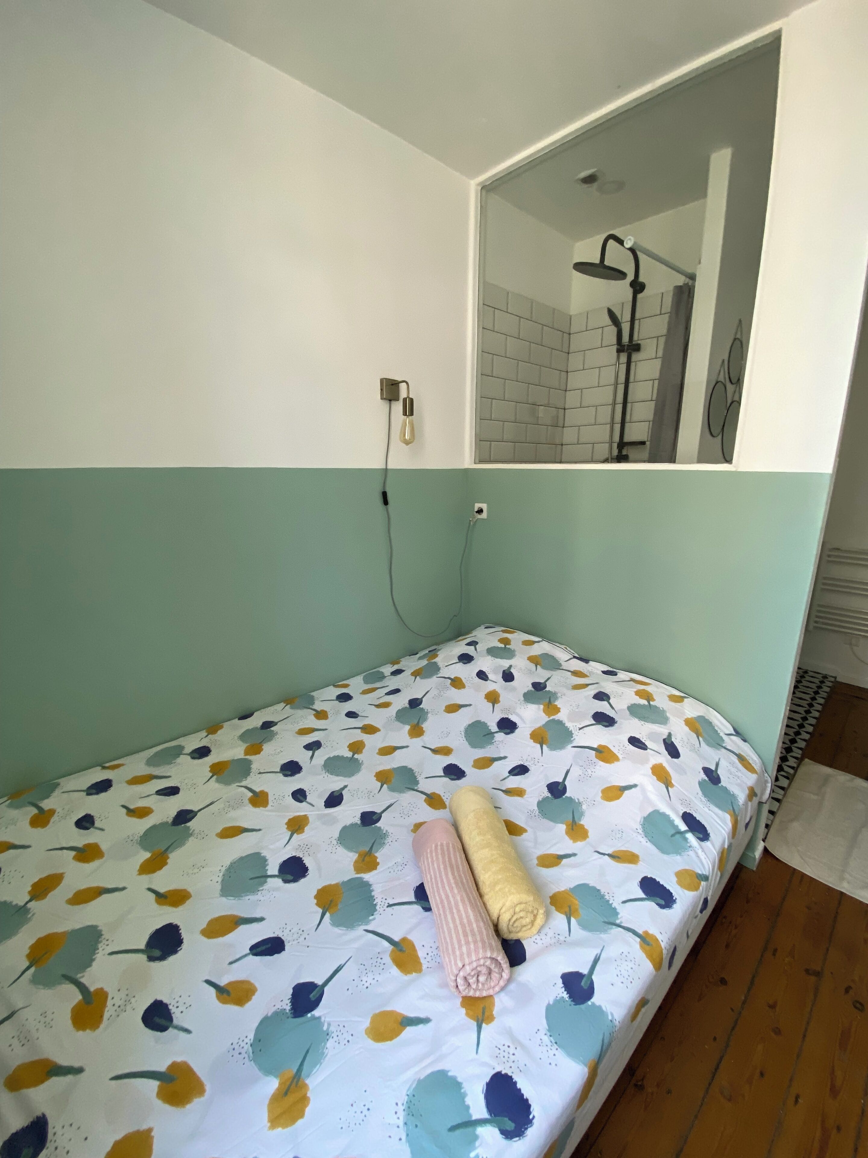 Een kleine eenpersoonskamer met een bedovertrek met een patroon, een spiegel boven het bed, en een zichtbare douche in de achtergrond.