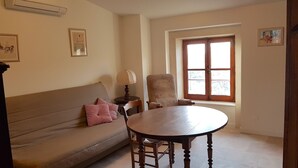 A living room with a sofa, a round wooden table, a chair, a lamp, and a window with a view of the outside. - Provence: Crossing city apartment, quiet, very bright, terrace. 2 stars. (VALREAS)