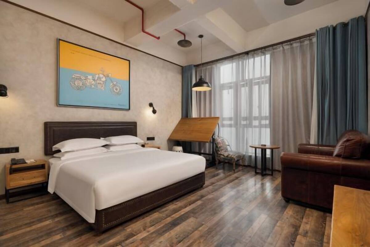 Habitación de hotel con una cama grande, piso de madera, escritorio, silla, sofá, un cuadro en la pared y una ventana con cortinas.