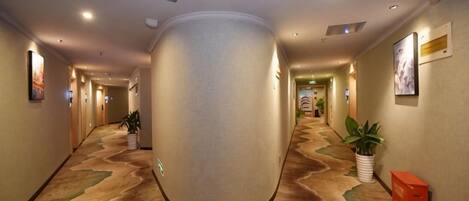 A hotel hallway with a central pillar, framed pictures on the walls, and a carpeted floor.