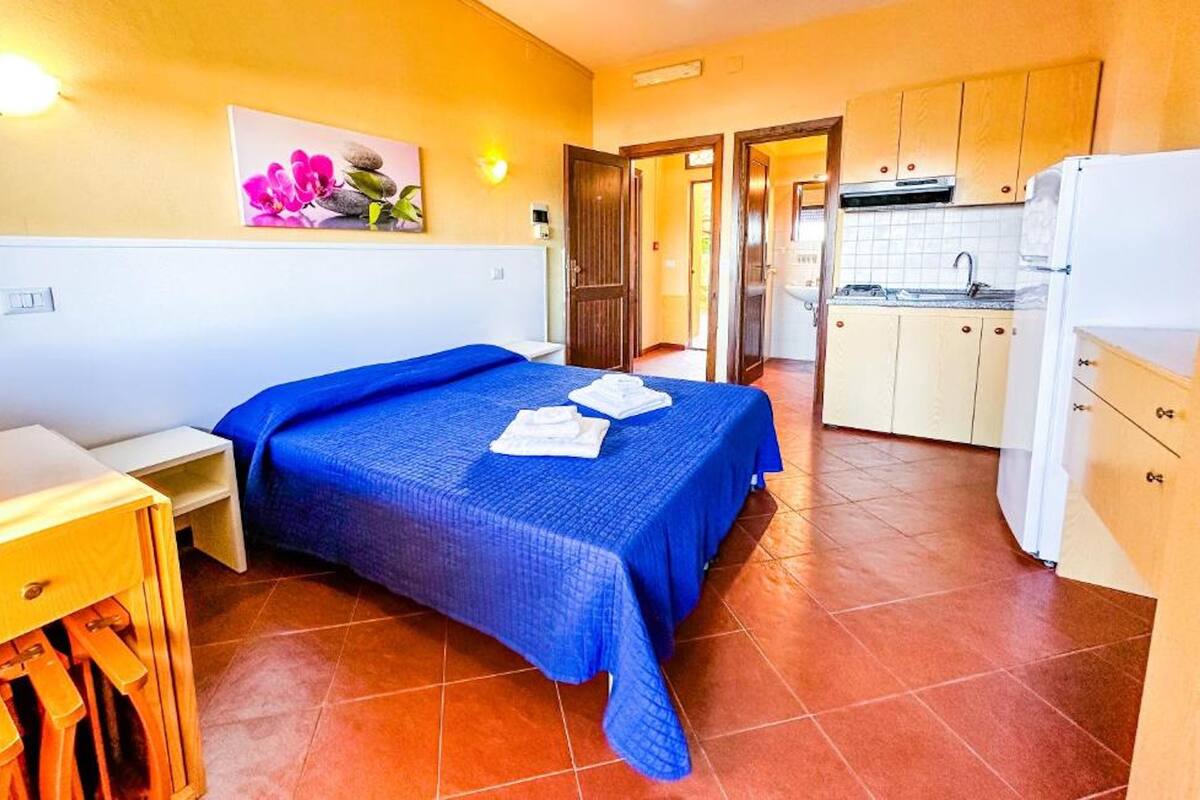 Une chambre avec un lit, une petite table et une table de chevet. Il y a également une kitchenette équipée d’un évier et d’un réfrigérateur.