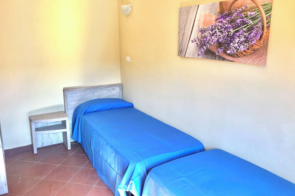 Une petite chambre avec un lit simple, une tête de lit en bois, un banc et un luminaire fixé au mur.