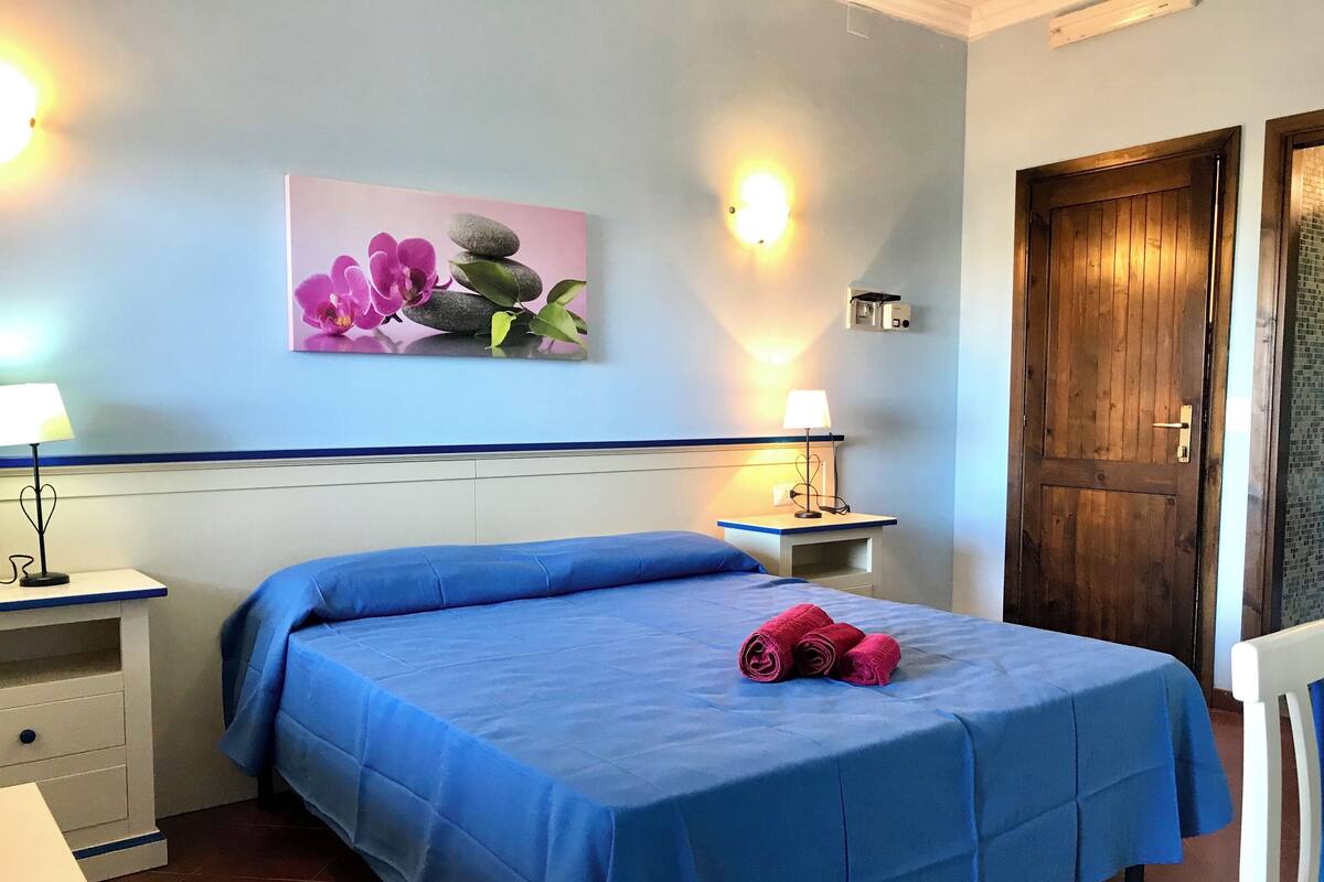Une chambre d’hôtel comprenant un lit, deux tables de chevet, une petite table, une porte et un luminaire fixé au mur.