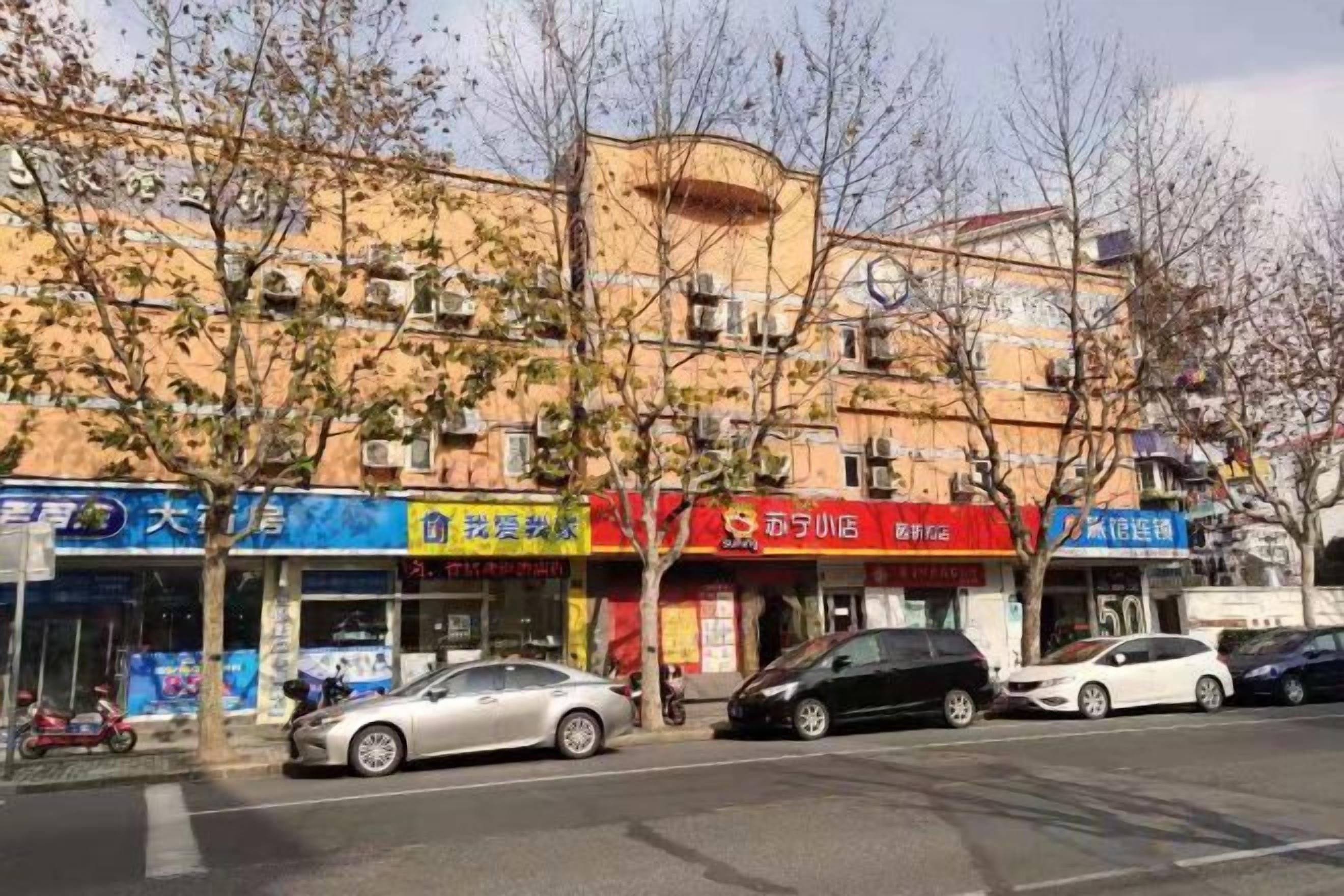 Straßenansicht mit mehreren Geschäften, darunter eine Apotheke, ein Restaurant und ein Kiosk. Entlang des Bürgersteigs stehen parkende Autos.