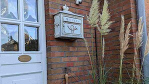 A white door with a window above it, a mailbox, a broom, and tall grasses against a brick wall. - Haus Hanne Sylt ♡ Holiday home for 6 people for families, 2 generations & dog (Sylt)