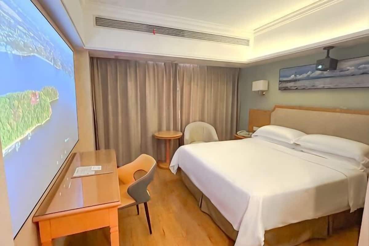 Hotellihuone, jossa on sänky, työpöytä, tuoli, pieni p öytä, seinällä taulu ja suuri näyttö, jossa näkyy kartta.