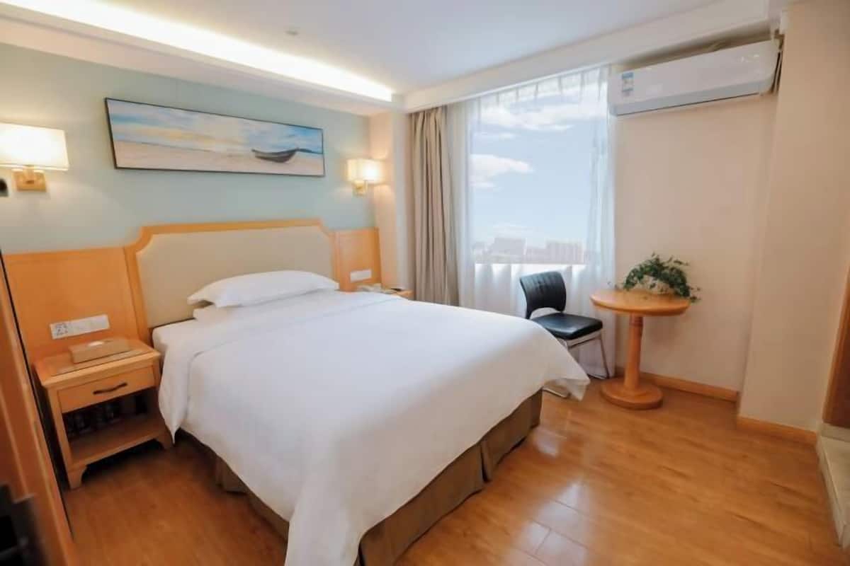Hotellihuone, jossa on suuri sänky, yöpöytä, lamppu ja pieni pyöreä pöytä, jonka päällä on kasvi.