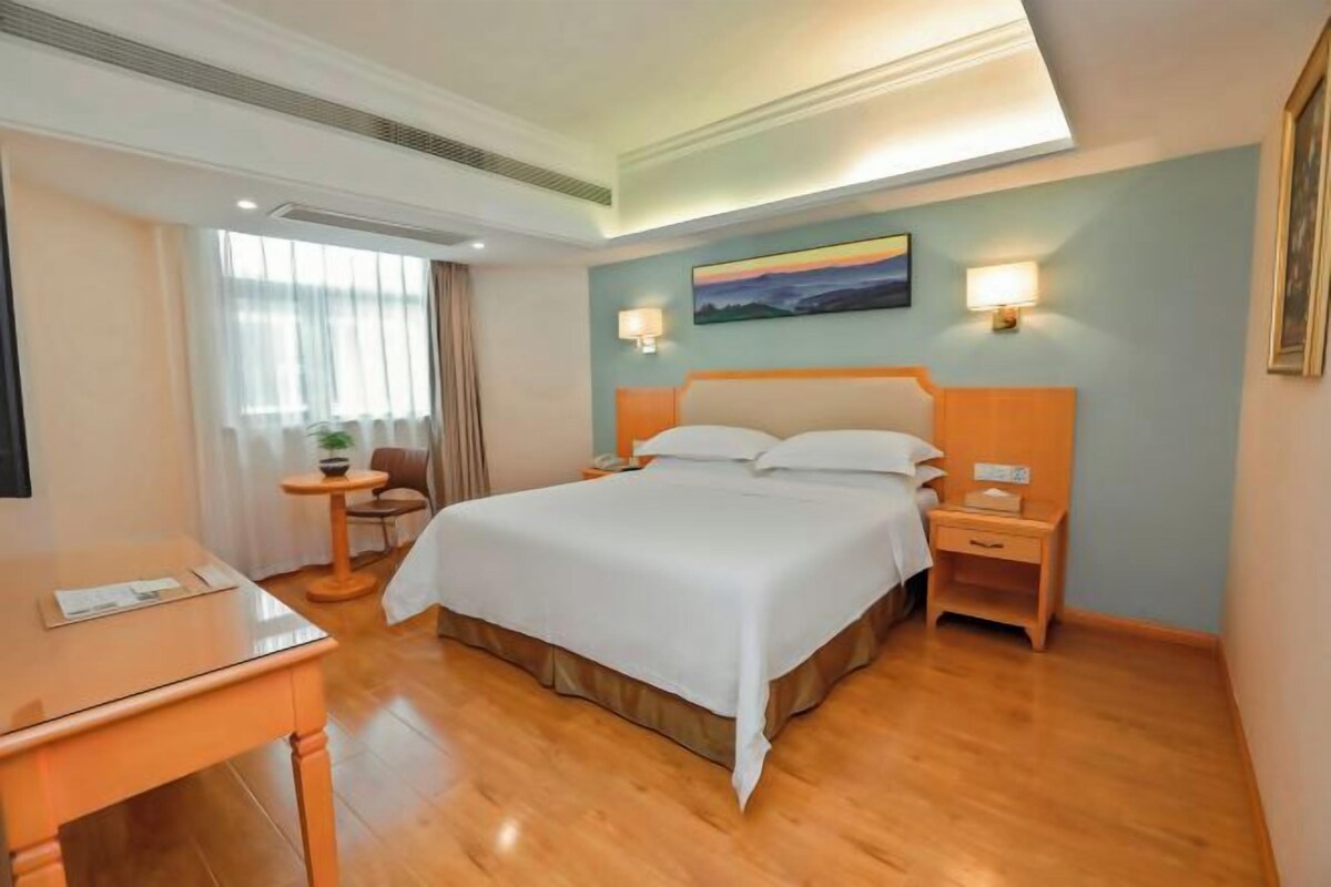 A hotel room with a bed, bedside tables, a desk, a chair, and a window with curtains.