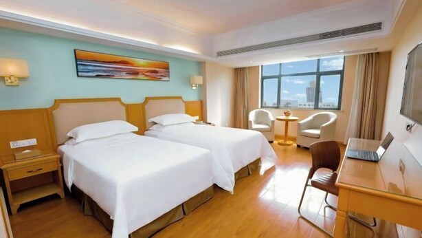 A hotel room with two beds, a desk with a laptop, a chair, and a view of the city. - Vienna 3 Best Hotel Jingmen Wanda (Jingmen)