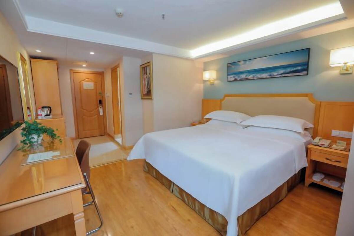 Quarto de hotel com cama, escrivaninha, cadeira, mesa de cabeceira, abajur e um quadro na parede.
