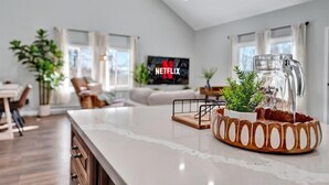 A modern living room with a large TV displaying the Netflix logo, a kitchen area with a marble countertop, and a dining area with a table and chairs.