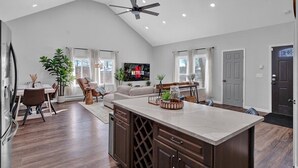 A spacious kitchen with a large island, a dining area with a table and chairs, a living room with a sofa, and a TV mounted on the wall.