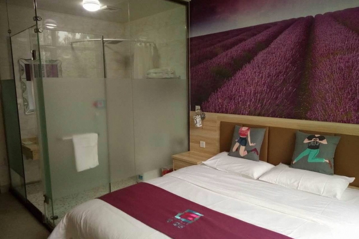 Quarto de hotel com chuveiro em cabine de vidro, cama com roupa de cama branca e painel de parede floral roxo.