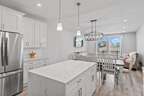 A modern kitchen with white cabinetry, a large island, stainless steel appliances, pendant lighting, and a dining area with a view of the outside.