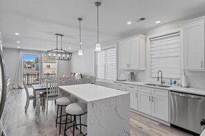 Una cocina moderna con armarios blancos, una isla de mármol, iluminación colgante y vistas al exterior.