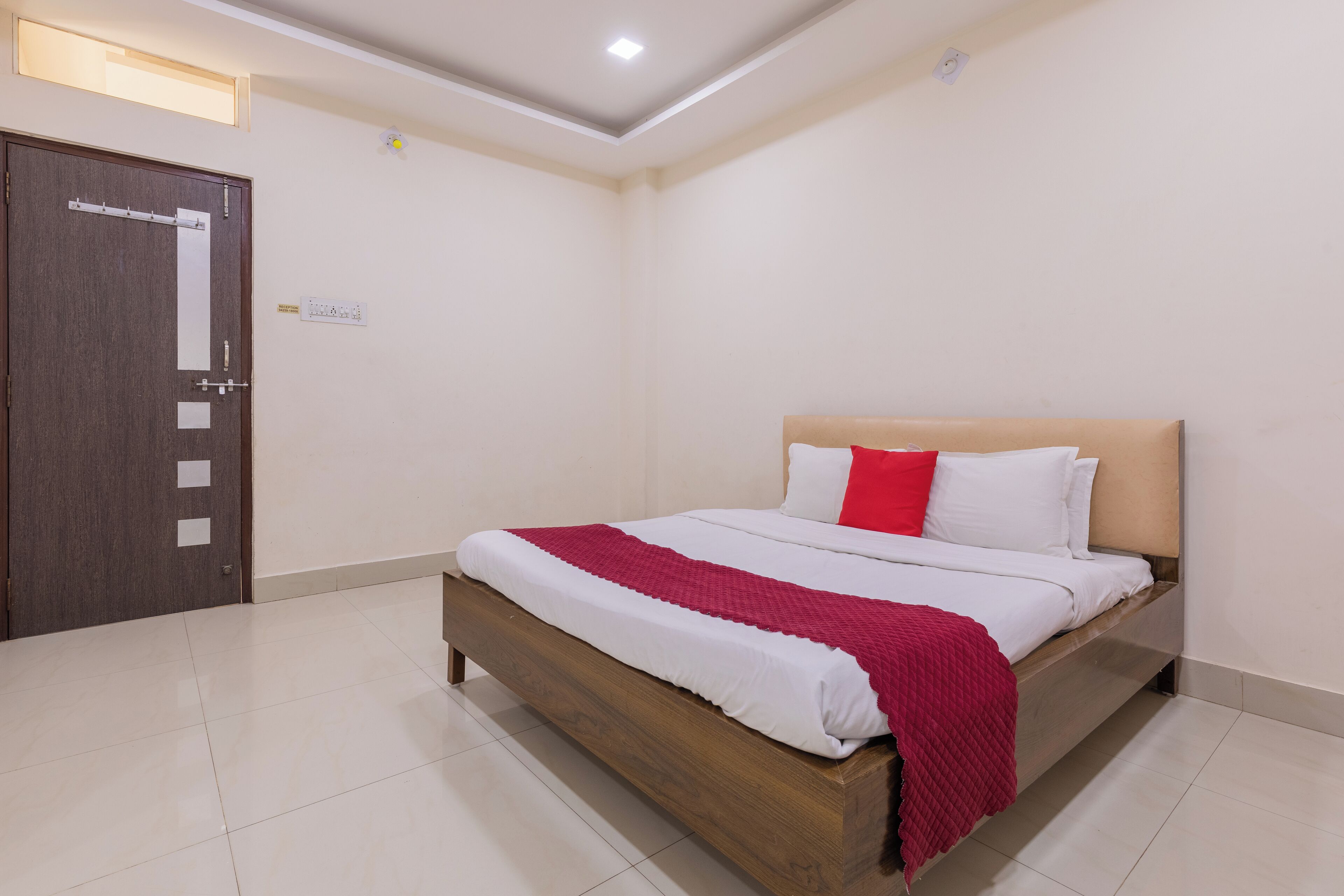 A bedroom with a wooden bed, white bedding, a red pillow, and a door with a light switch.
