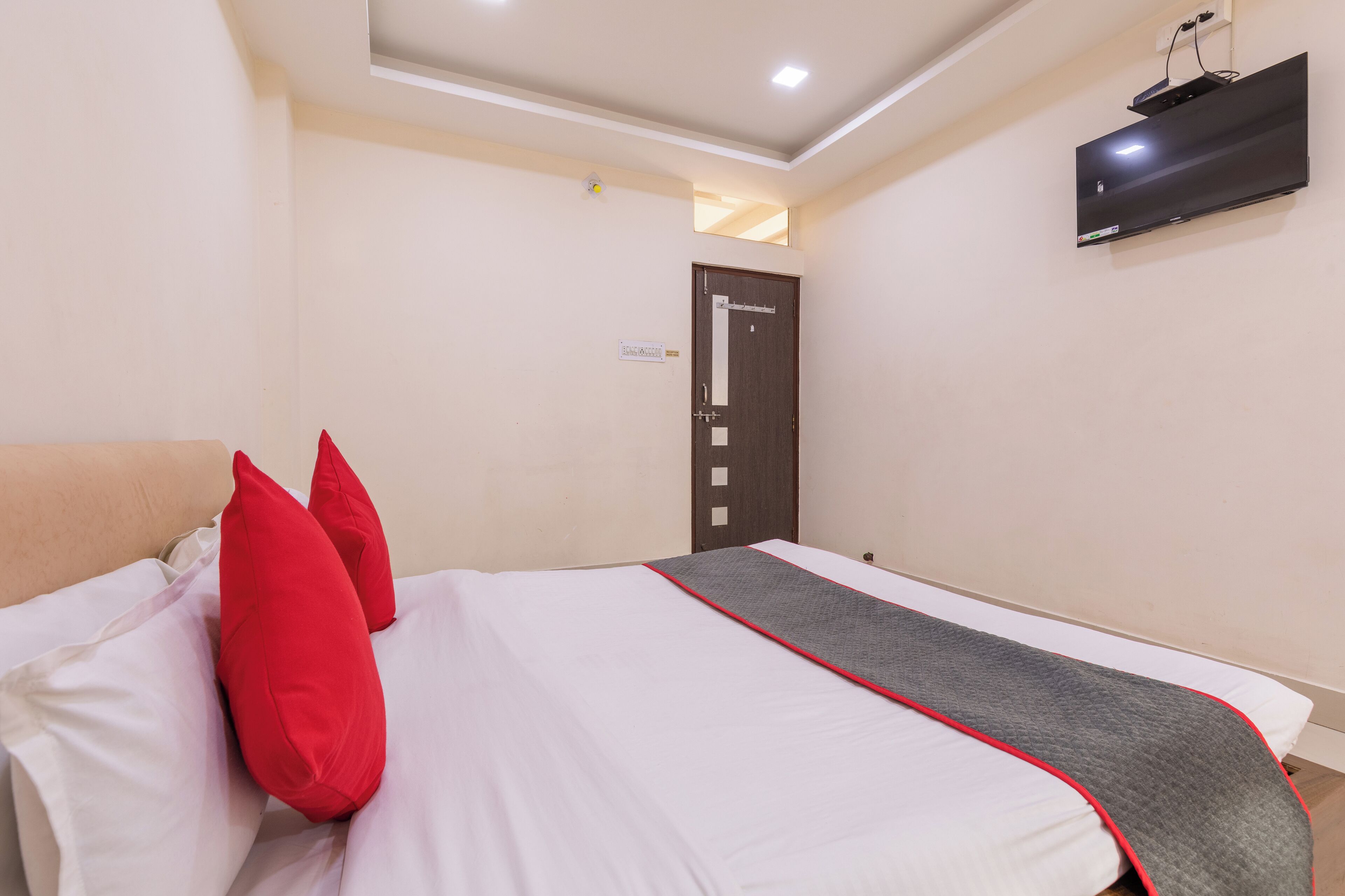 A hotel room with a bed, red pillows, a grey blanket, a TV mounted on the wall, and a door with a sign.